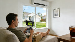 Lounging on a sofa, a man uses an attached FPV screen controller to guide his Mowrator mower through the window view