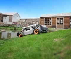 A close-up view of the Mowrator cutting lush, steep grass near a water feature, with rustic wooden and white buildings in the background.