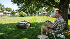 A man relaxes under a tree, using a remote control to operate his Mowrator lawn mower across a sunny backyard