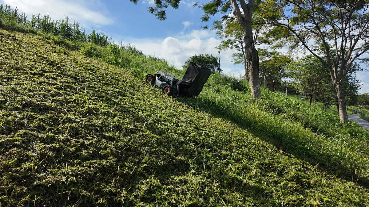 Mowrator remote control lawn mower with collection bag ascending steep grassy slope near trees under blue sky