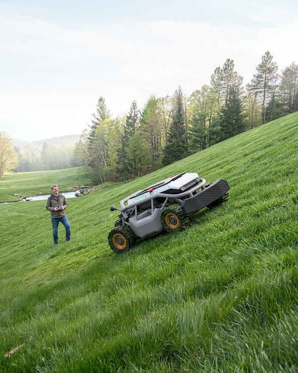 A man stands on a lush green hill, using a remote control to operate a Mowrator slope mower on a steep incline