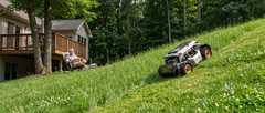 Robotic tracked lawn mower with orange wheels climbing a grassy hill in a backyard while a man watches from the deck