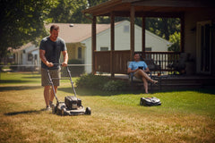 Exhausted person pushing a lawn mower in intense summer heat haze contrasted with a person using a remote control mower from a shaded porch