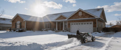 A Mowrator unit equipped with a snow plow clears a snow-covered driveway in front of a house on a sunny winter day