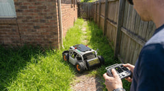 A remote operator guides the Mowrator mower down a tight, grassy passage between a brick house and a wooden fence