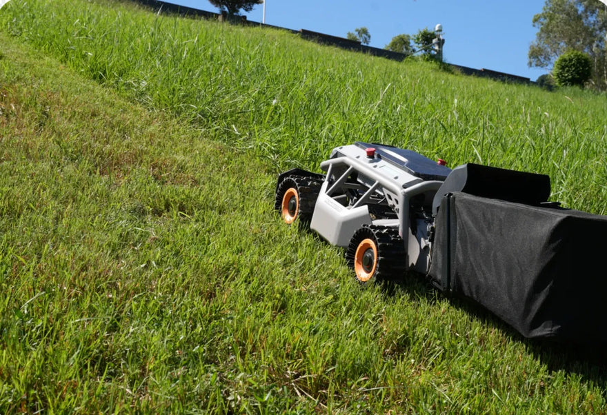 Mowrator robotic mower with black collection bag and orange wheels climbs green grassy hillside