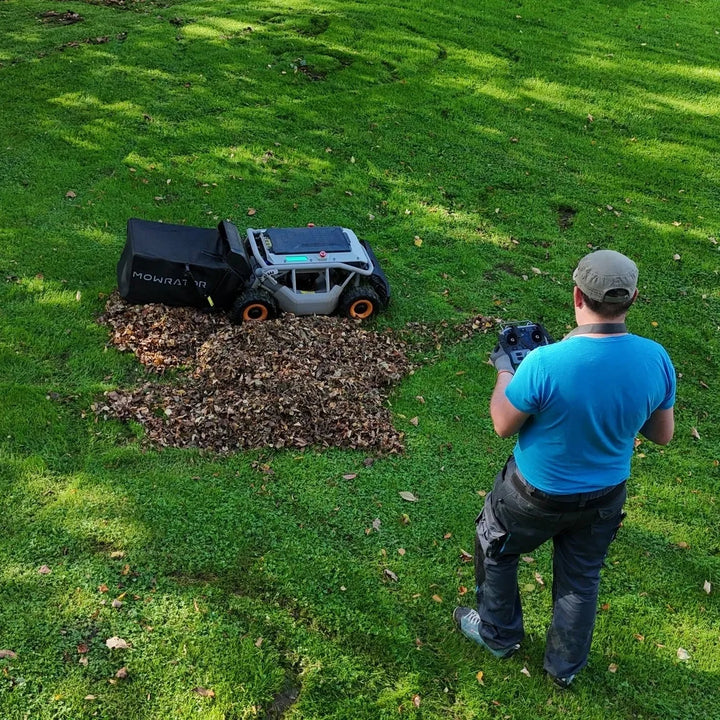 Mowrator remote-controlled leaf vacuum collects pile of brown leaves on sunny grassy lawn with operator holding remote